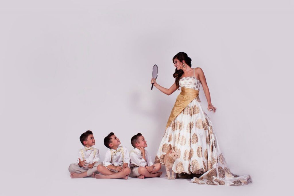 Woman in gold and white gown holding mirror, three boys seated look up at her. White background.
