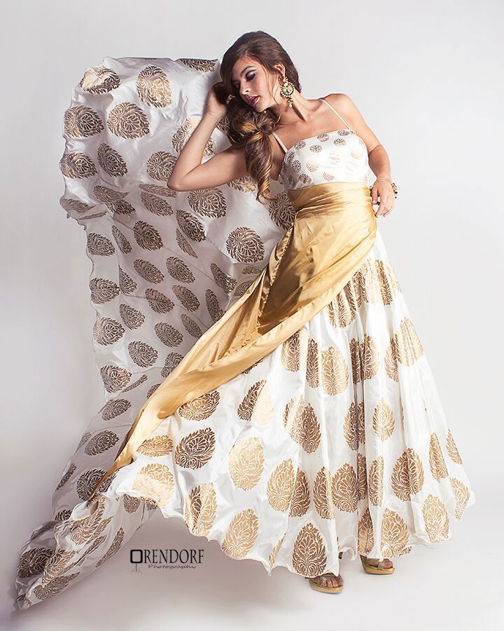 Woman in a white and gold patterned formal gown, holding flowing fabric against a white backdrop.
