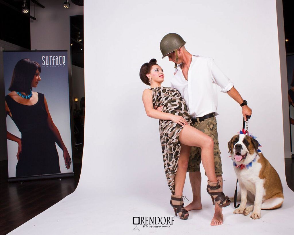 Man in helmet and woman pose with a large dog in a studio setting. Advertisement banner in background.