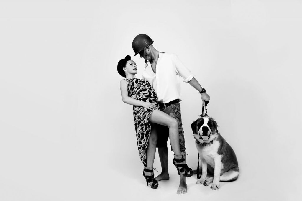 Couple dancing next to a Saint Bernard dog, man in a helmet and woman in a patterned dress, all against a white backdrop.