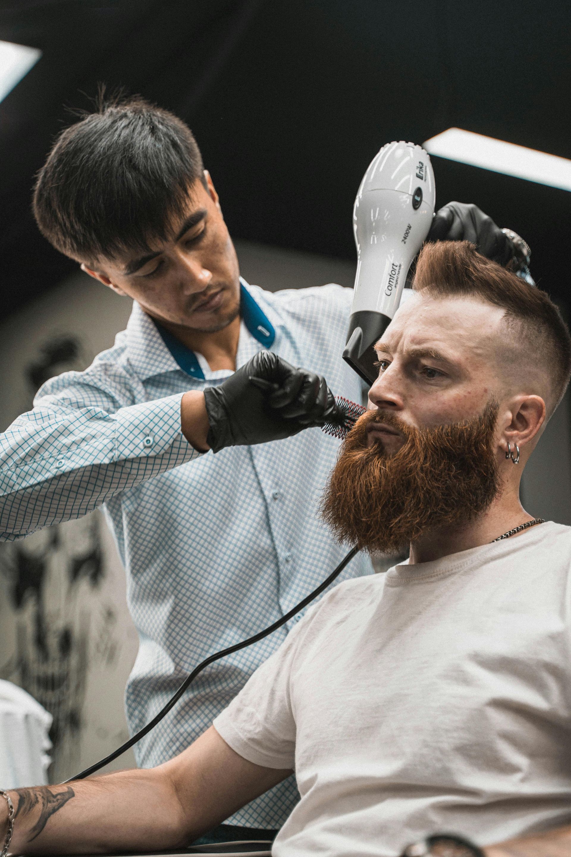 Barber trimming a client's beard in a barbershop. Barber wears gloves, holds clippers and a hairdryer. Client has a red beard.