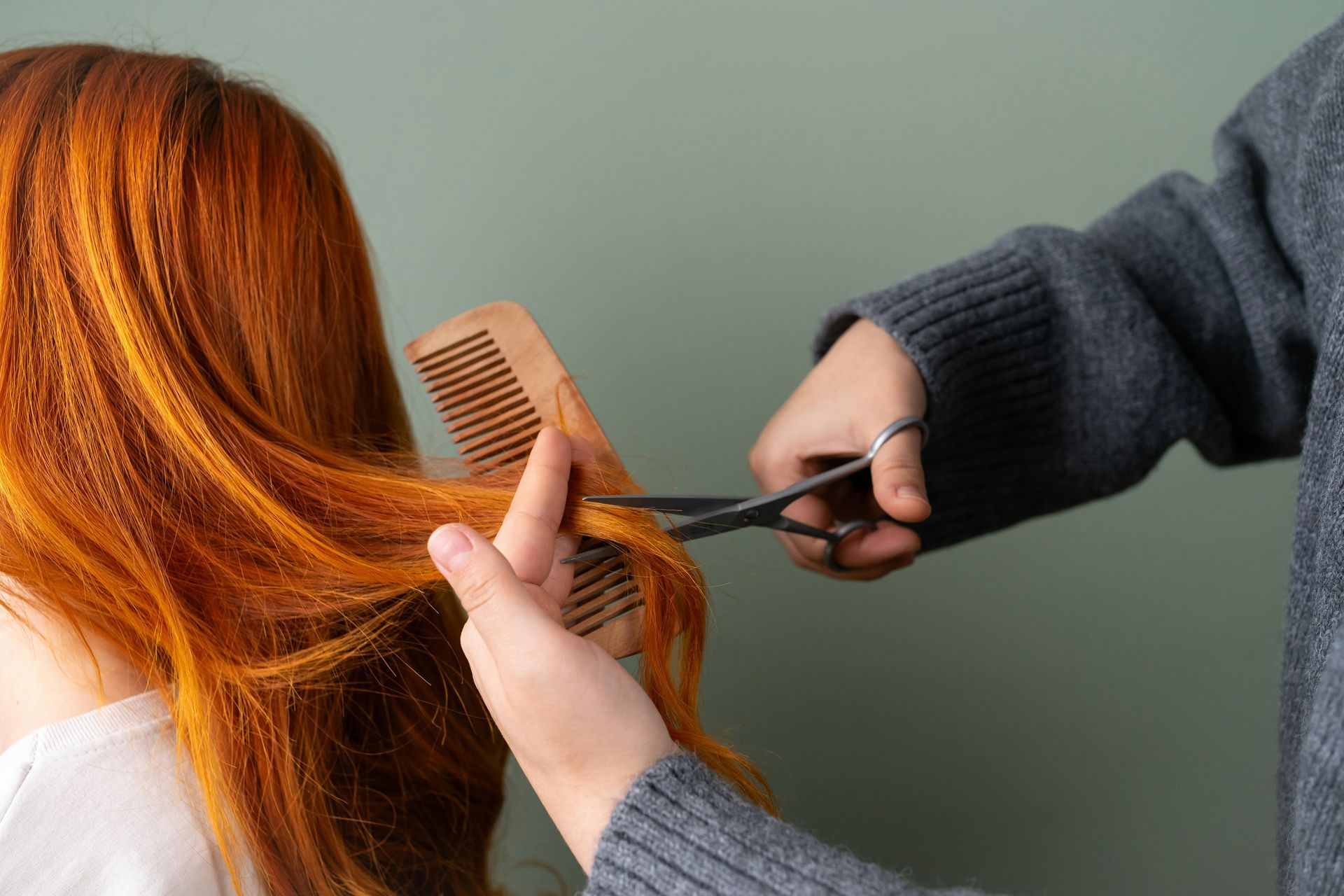 Stylist cutting long red hair with scissors while holding a comb, close-up against a plain green background