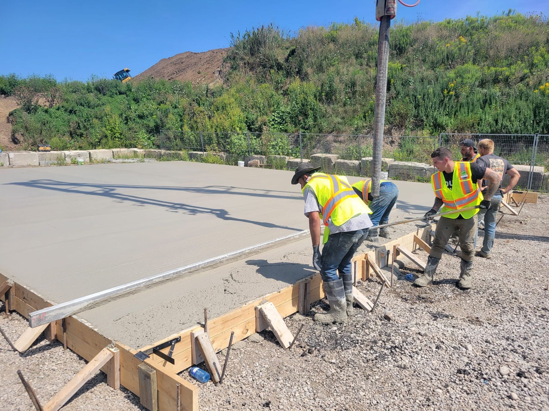 Workers pouring and leveling concrete on a roadbed beside a hillside under construction.