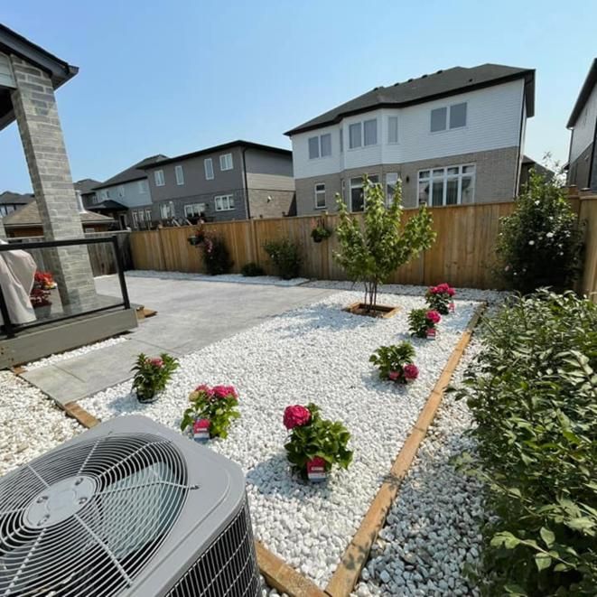 Backyard gravel garden with pink flowers, small trees, wooden fence, and a house air conditioner unit