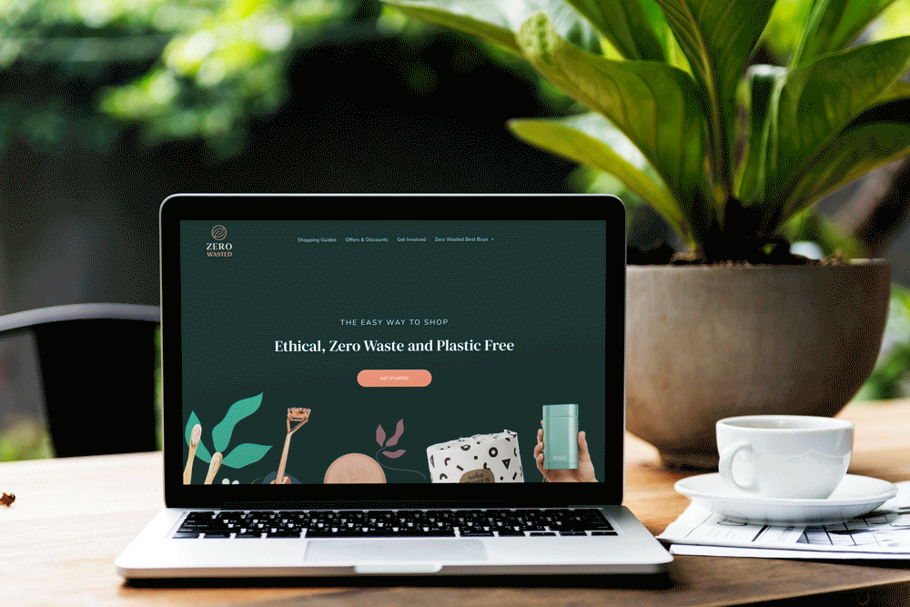 Laptop displaying website, coffee cup, and plant on a wooden table outdoors.