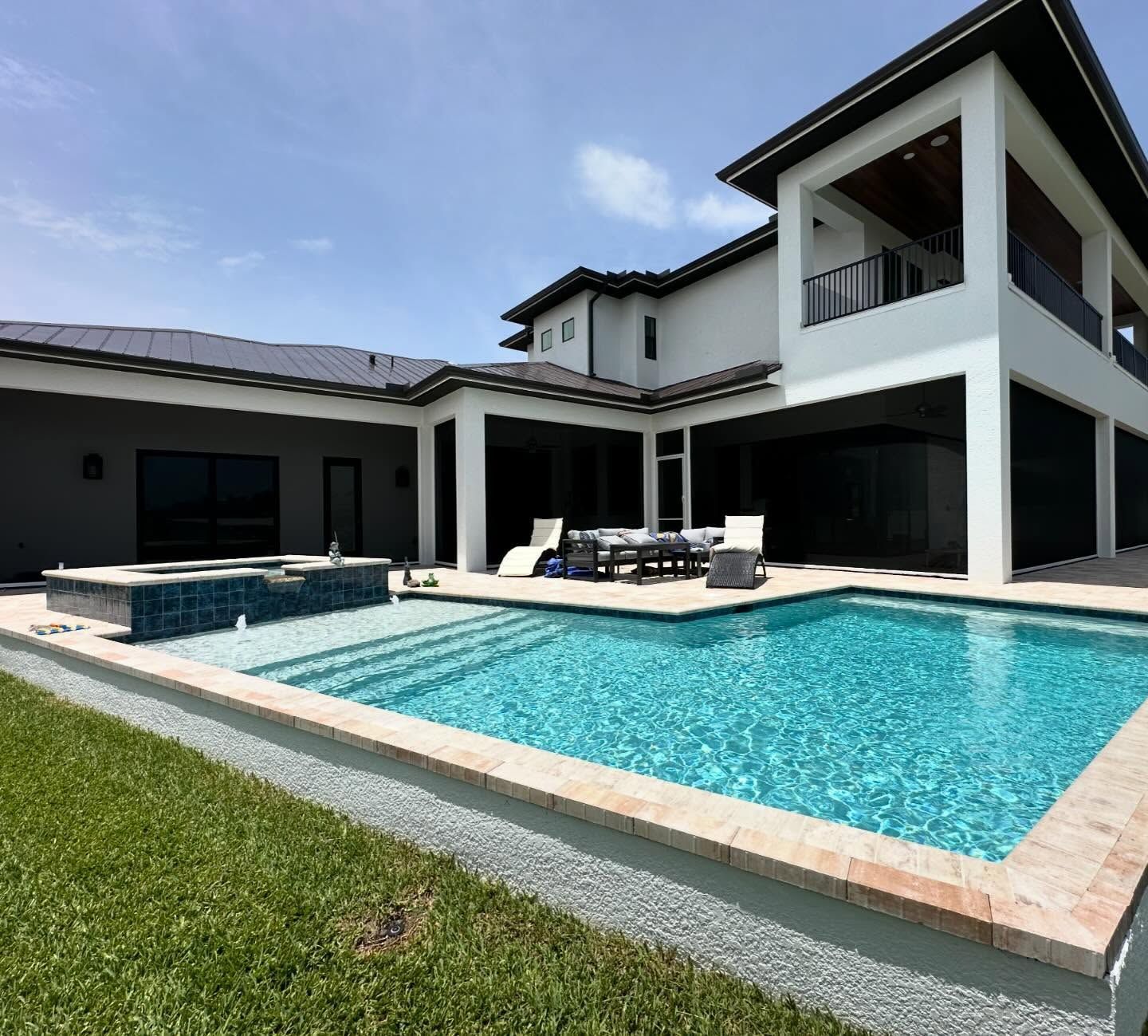 Modern house with a turquoise swimming pool, patio seating, and a green lawn under a blue sky
