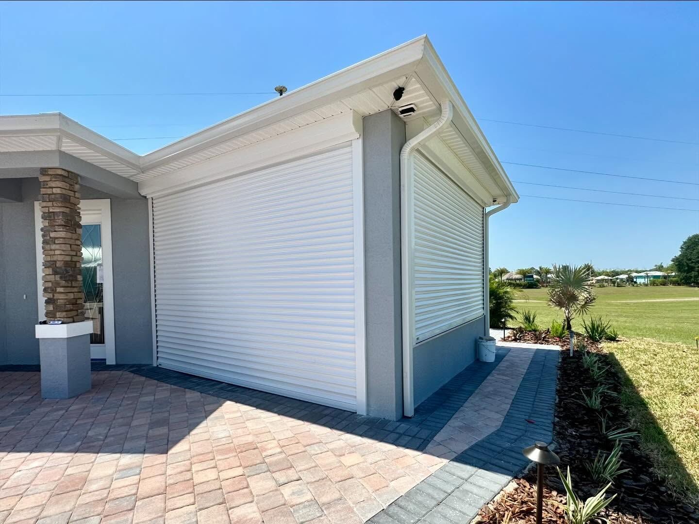 White house exterior with closed shutters, brick patio, and grassy field under a blue sky