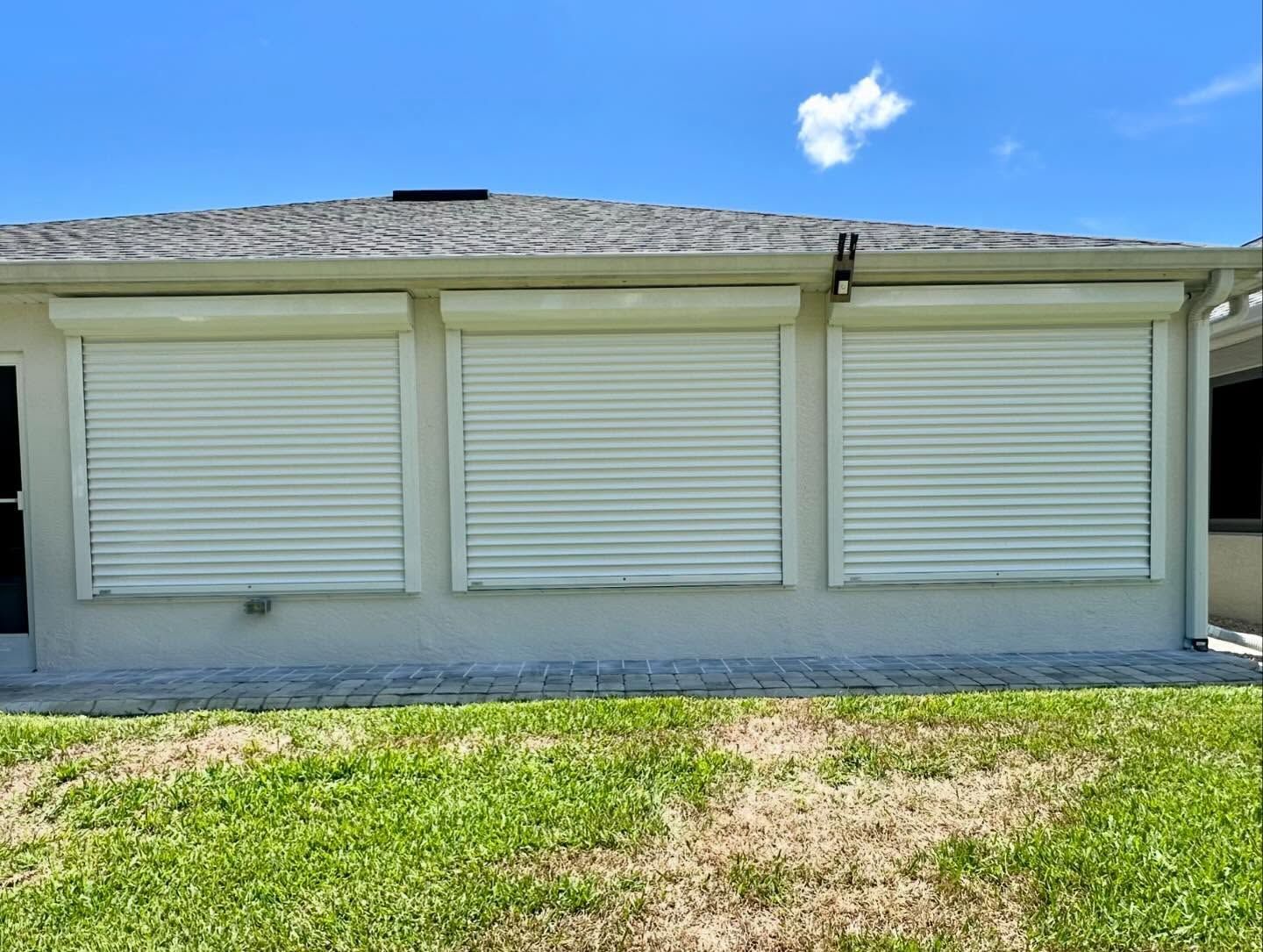 Single-story house exterior with closed white shutters and a grassy yard under a blue sky