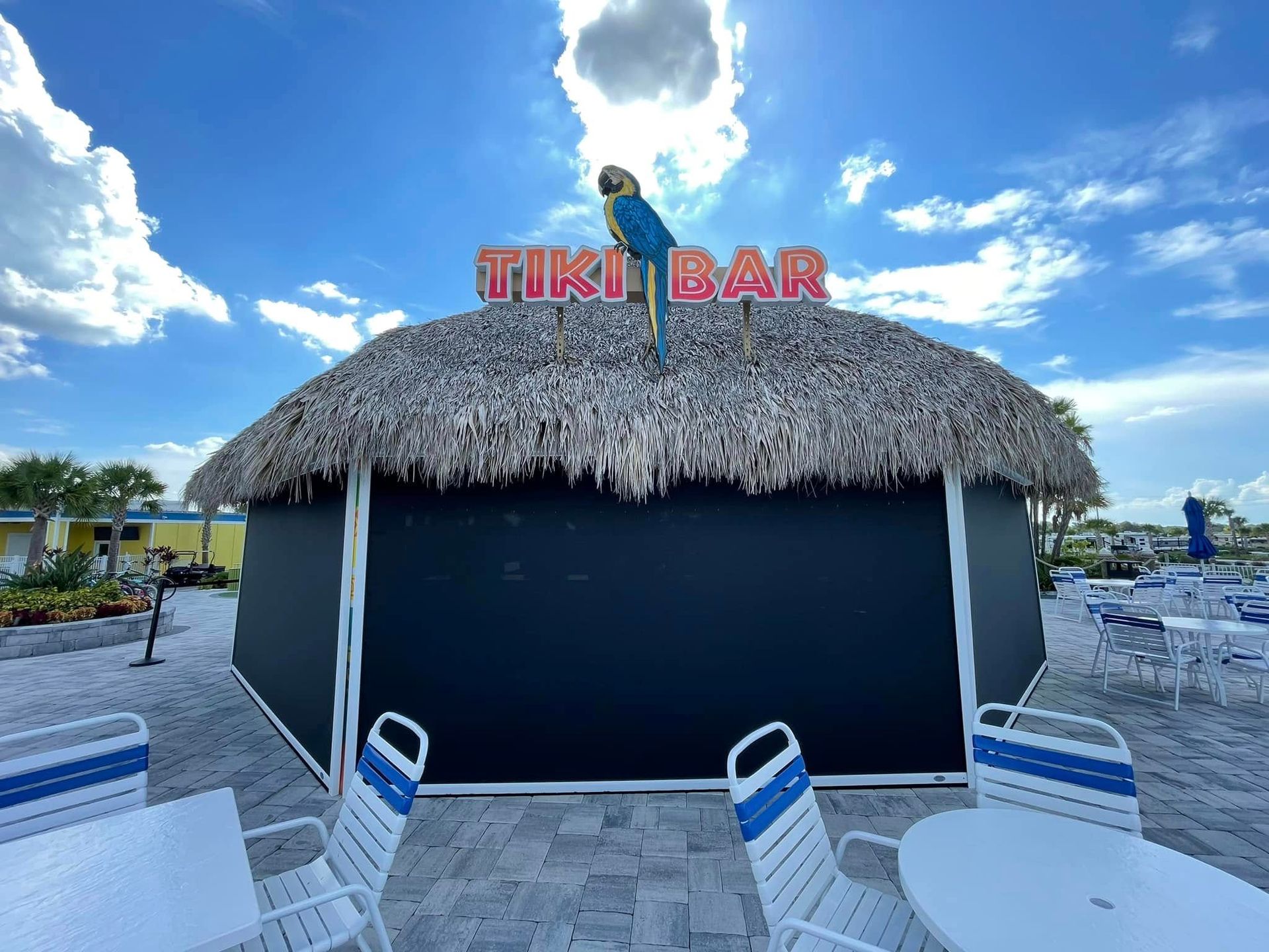 Thatched tiki bar with “TIKI BAR” sign, blue walls, and a paved seating area under a partly cloudy sky