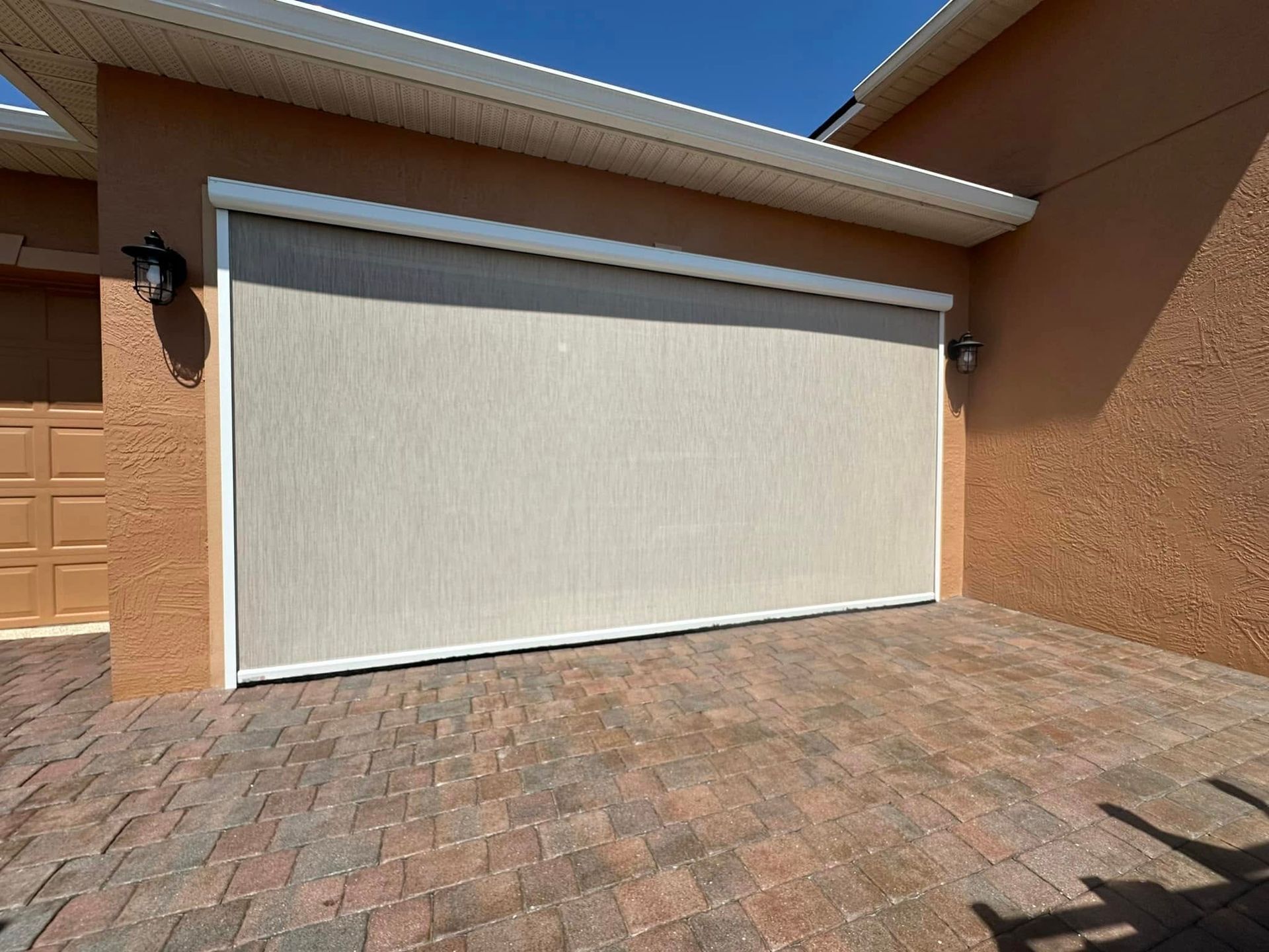Closed beige garage door on a stucco house with a brick-paved driveway in bright sunlight