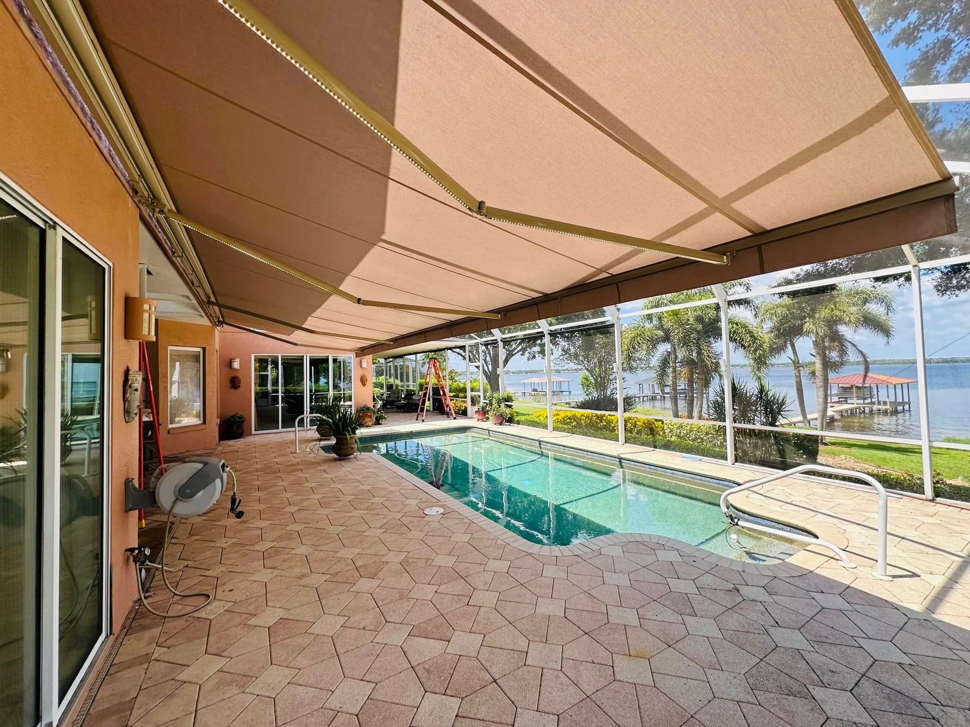 Covered patio with a long pool, tan tile floor, and ocean view beyond palm trees