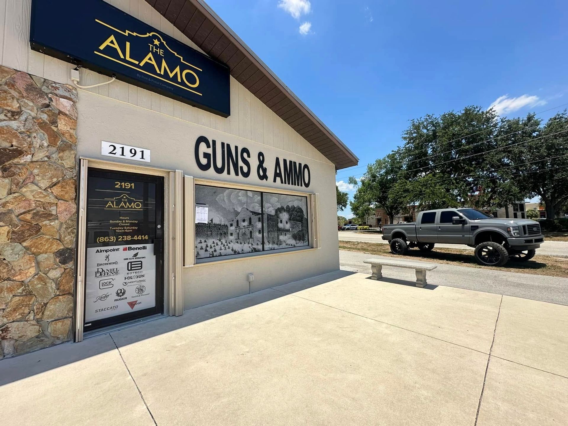 Alamo Guns & Ammo storefront with stone facade, large windows, and a black pickup in the parking lot