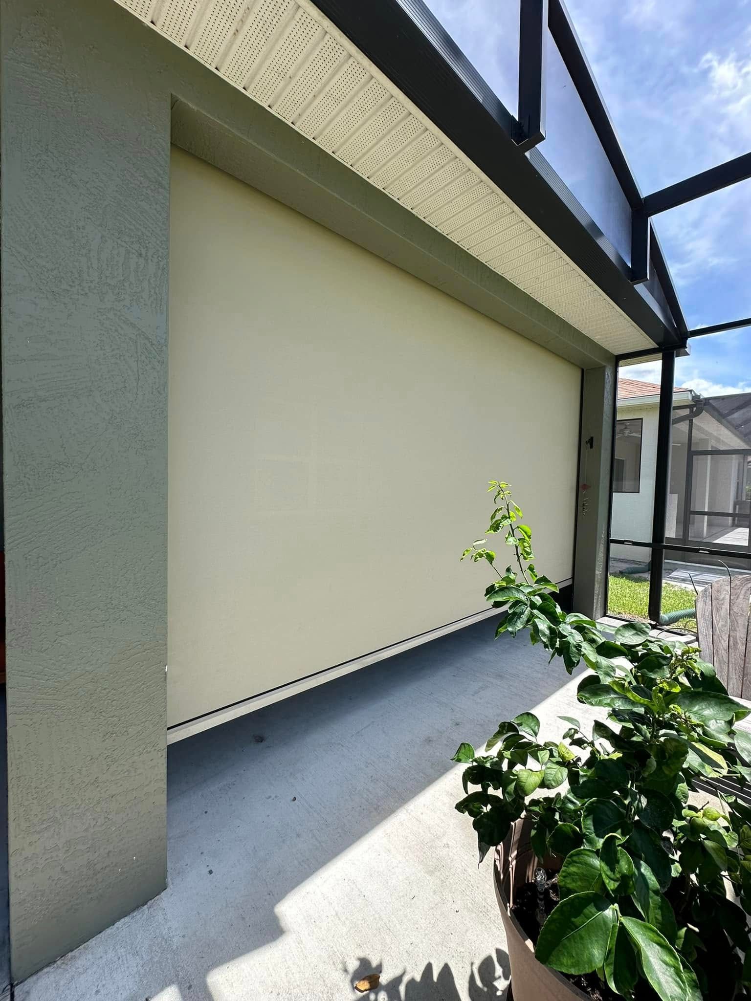 Covered outdoor patio with a closed cream roller blind, gray walls, and a potted green plant in sunlight
