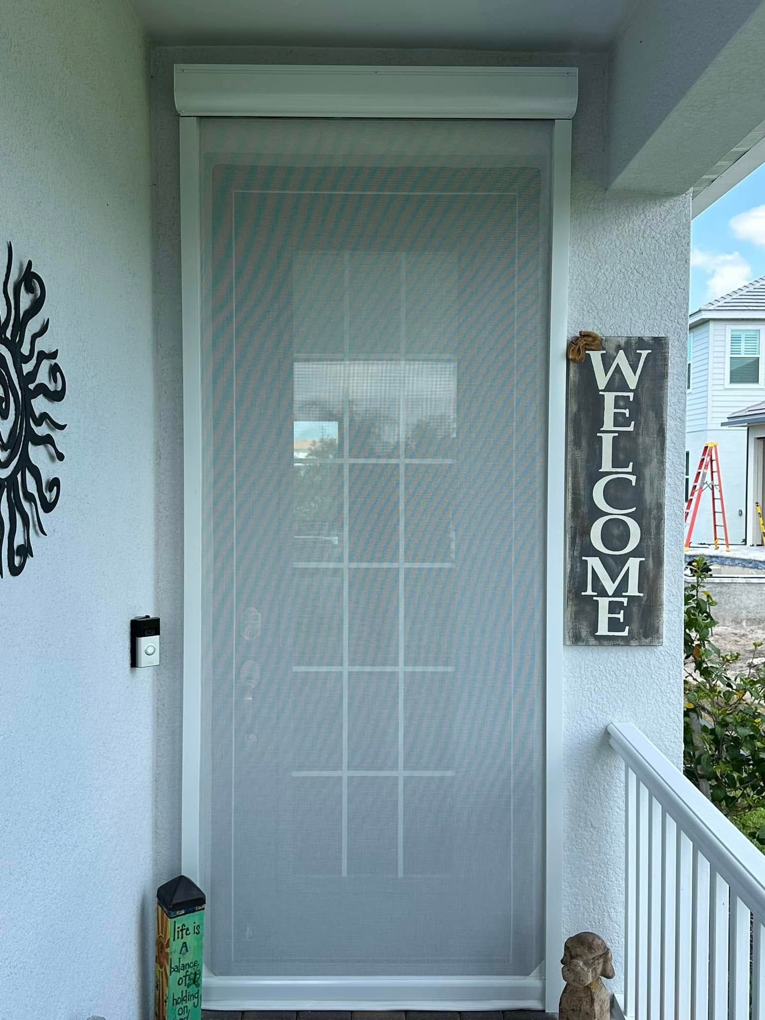Front porch with a screened door, vertical “WELCOME” sign, and railing beside a house entry.