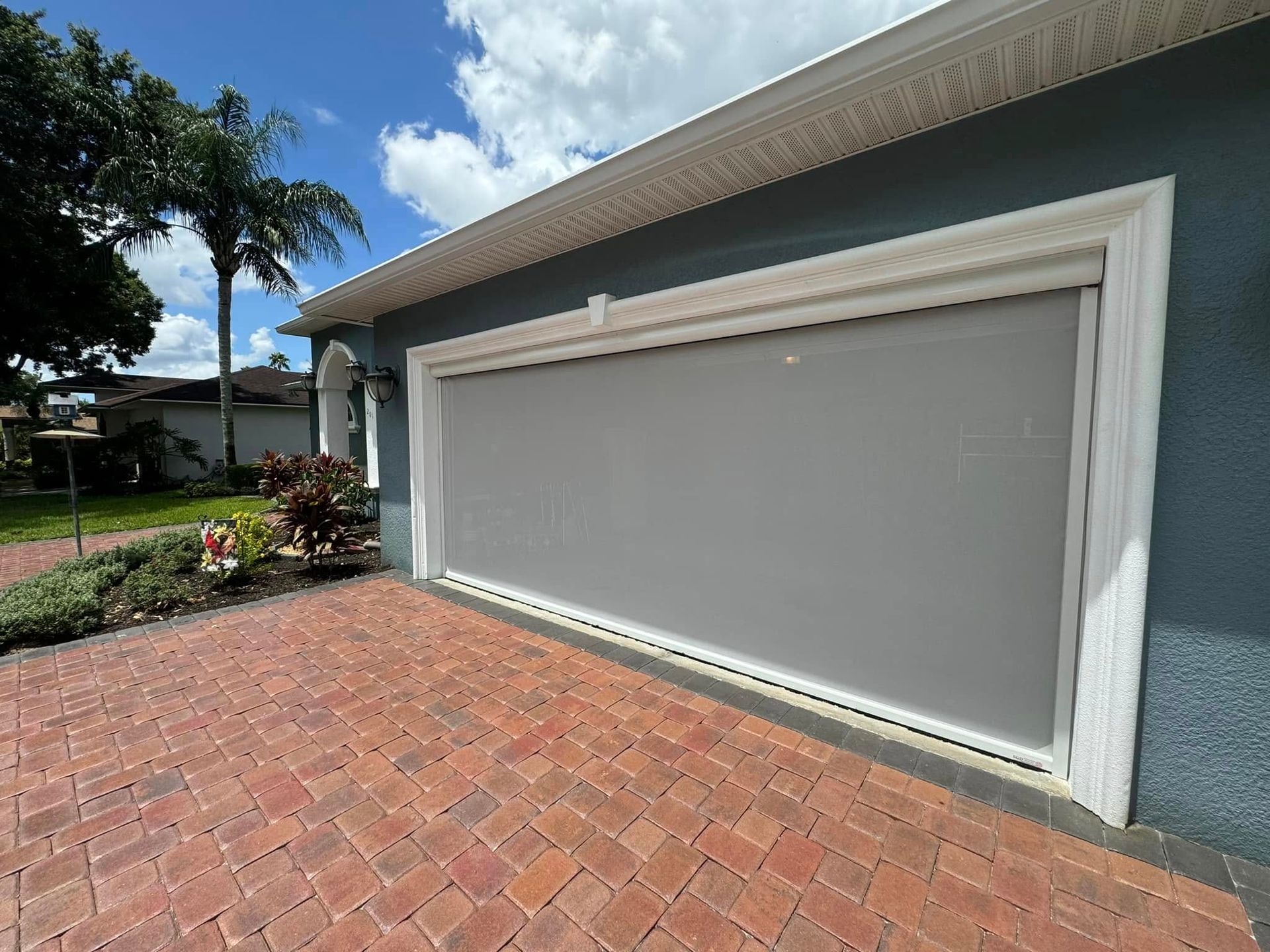 Closed white garage door on a gray house with a brick driveway and landscaping.