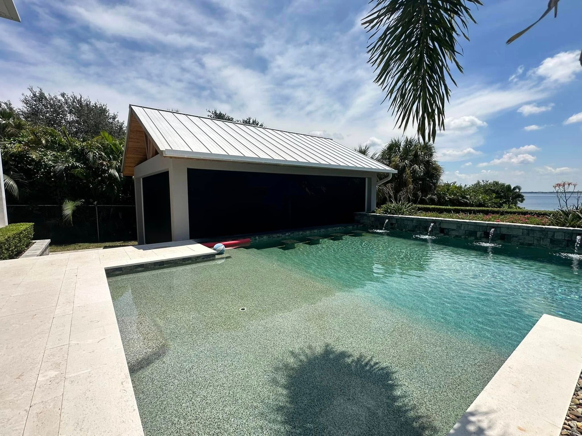 Sunlit backyard pool with a shallow beach entry, shaded cabana, and palm trees under a blue sky