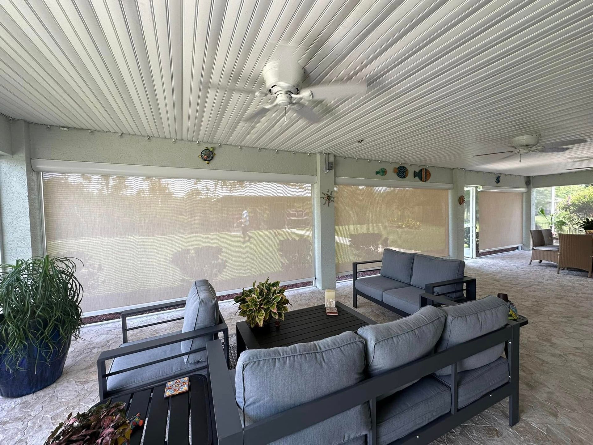 Covered patio with gray sectional seating, potted plants, and ceiling fans overlooking a yard