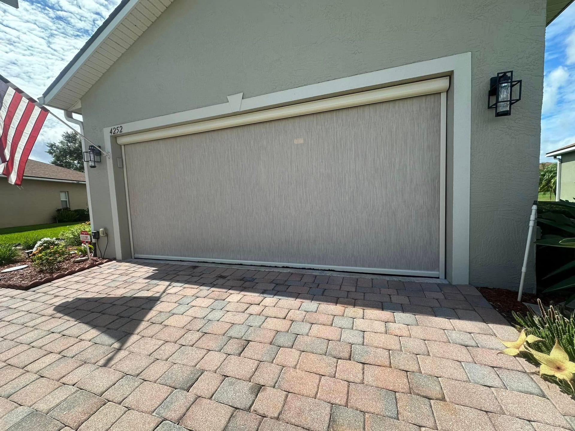 Closed garage door on a gray house beside a brick driveway and U.S. flag.