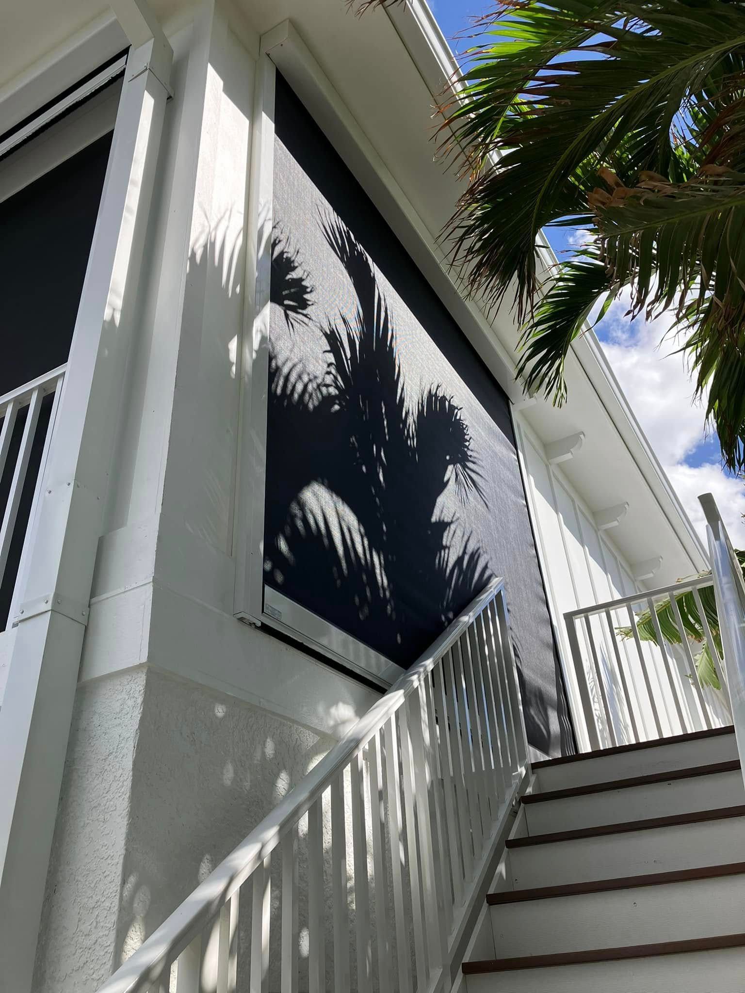 Stairway beside a white house with a dark window, shaded by palm leaves and bright sky