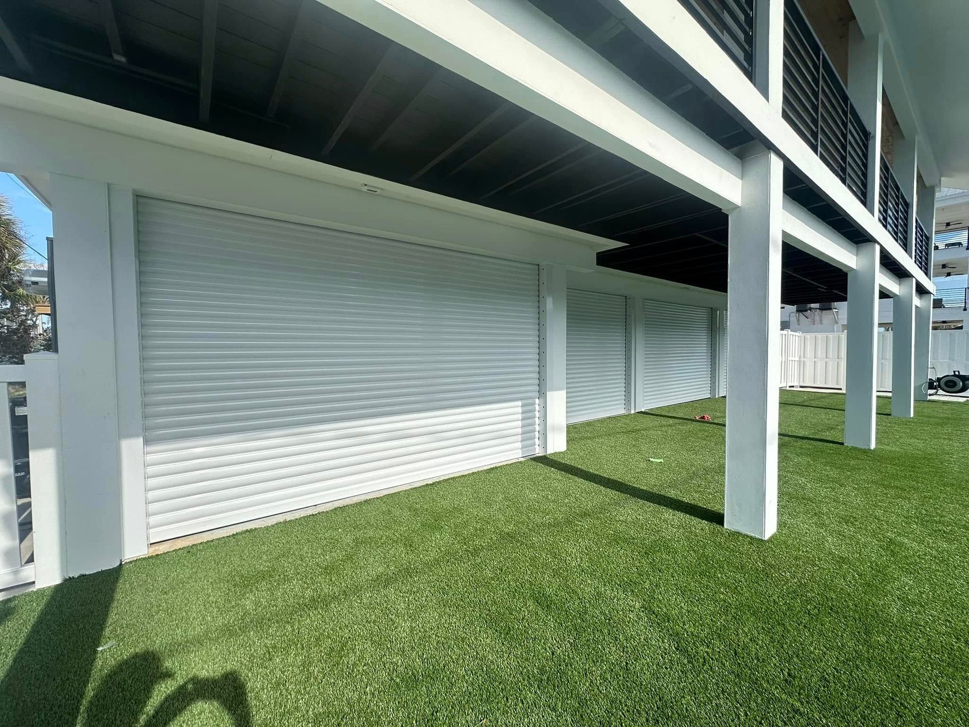 White building with closed roll-down shutters under a raised walkway, beside a grassy lawn