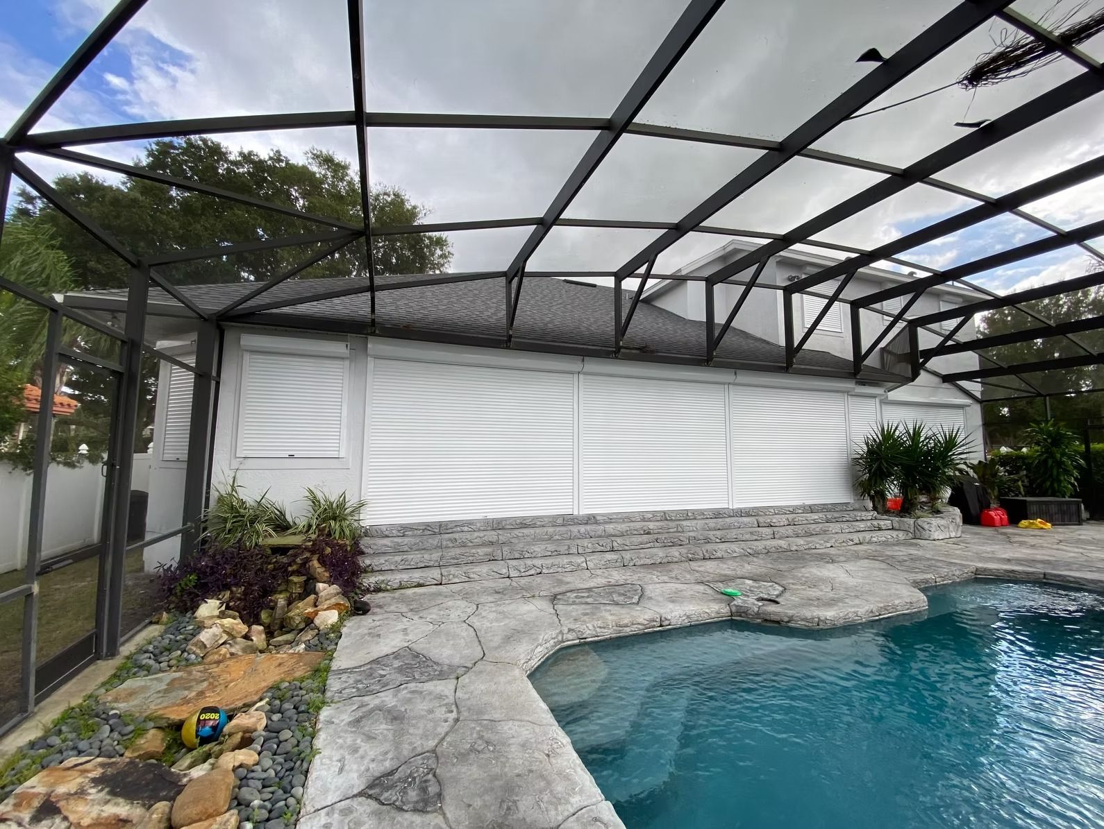 Pool area with a white shuttered house under a black-framed screen enclosure.