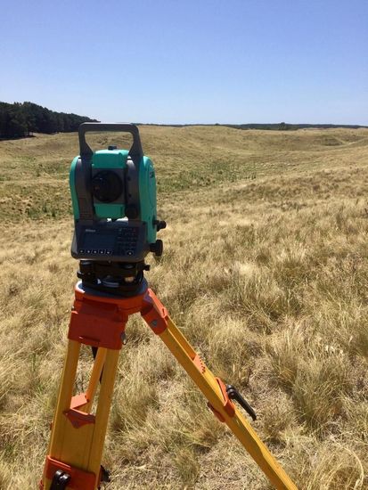 Instrumento de topografía sobre trípode en un campo de hierba bajo un cielo azul.