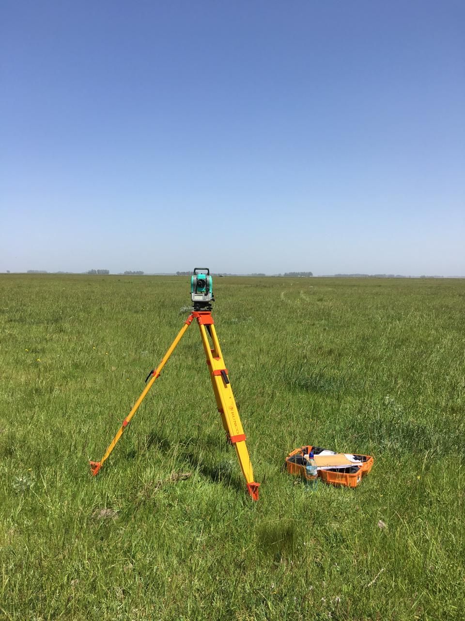 Trípode de topografía con instrumento en un campo de hierba bajo un cielo azul despejado.
