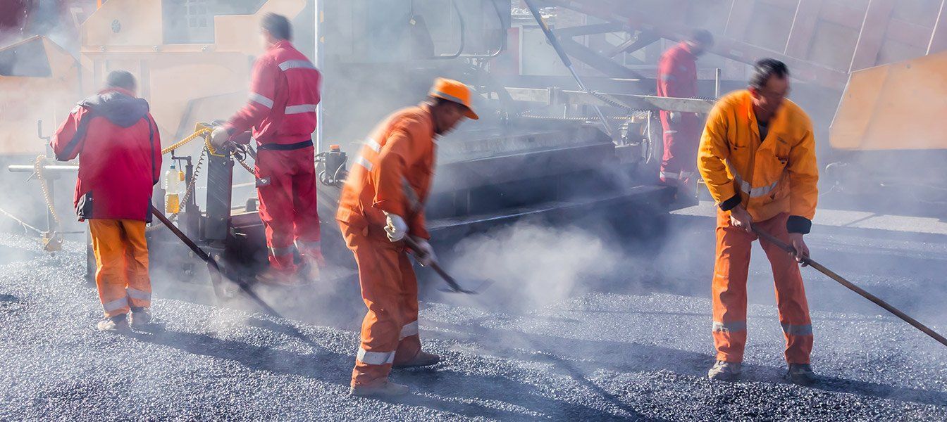Road workers in orange coveralls, using tools on asphalt. Steam rises from the work site.