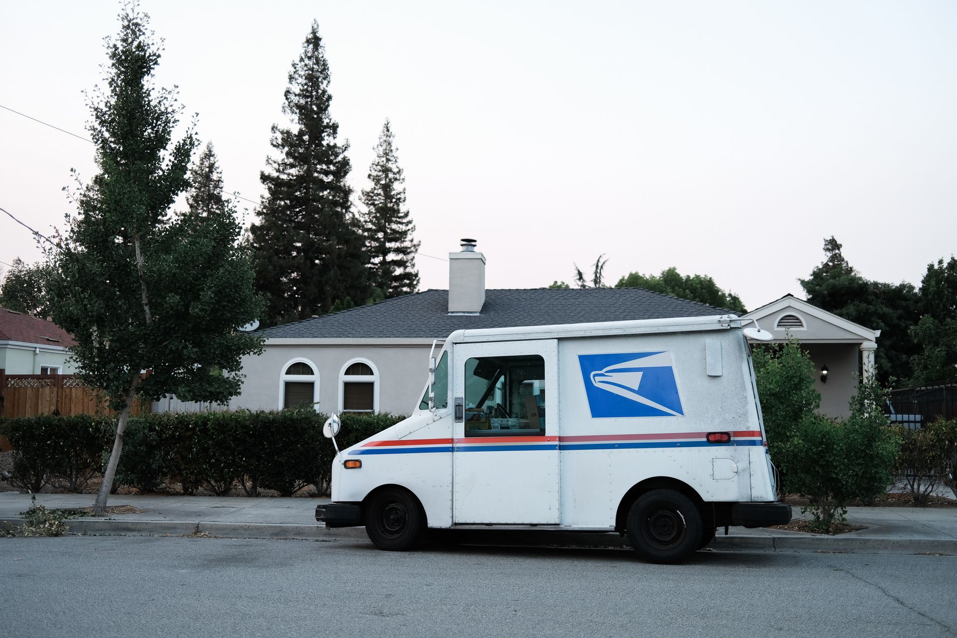White USPS mail truck parked on a residential street. Blue postal service logo on side.