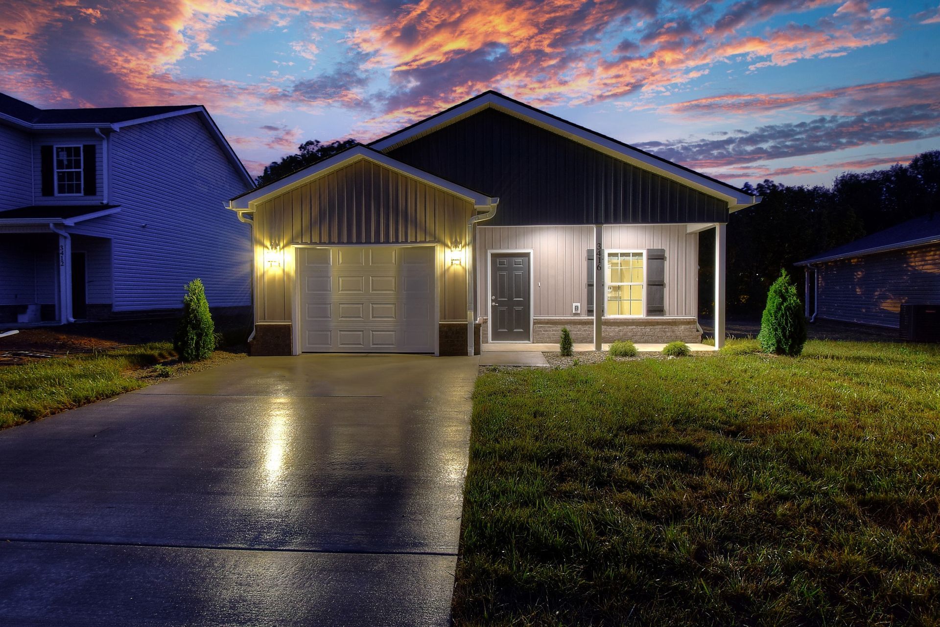 Suburban house at dusk with lit garage and porch, wet driveway reflecting the lights, and a colorful sunset sky