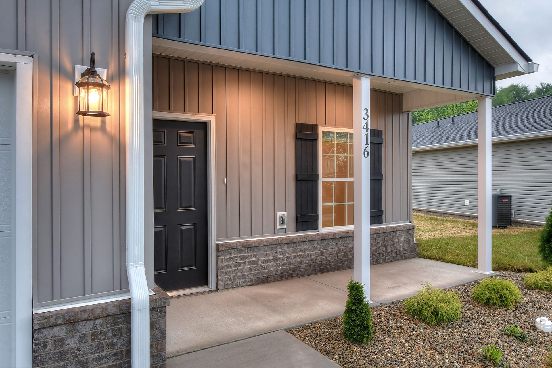 Front porch of a gray house with a black door, window, and lit wall lantern.