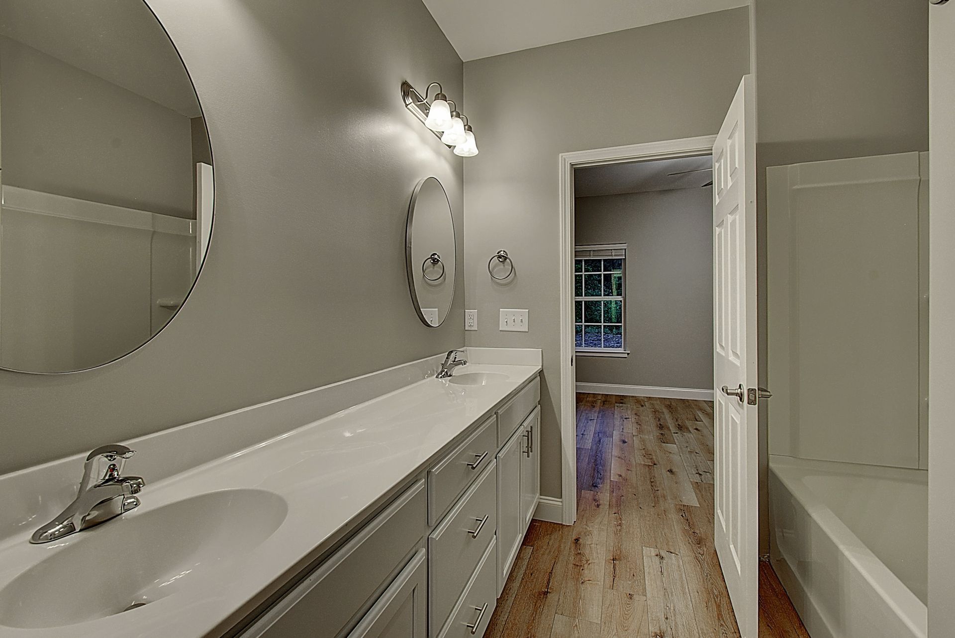 Bright bathroom with long vanity, mirror, tub, and open door to a bedroom beyond.