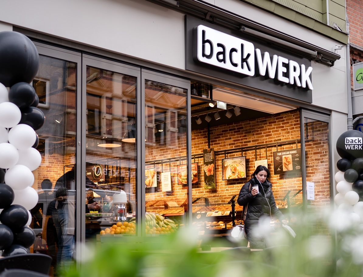 Bakkerijpui met backWERK-bord, warme verlichting en een persoon die buiten staat bij ballonnen en bloemen.