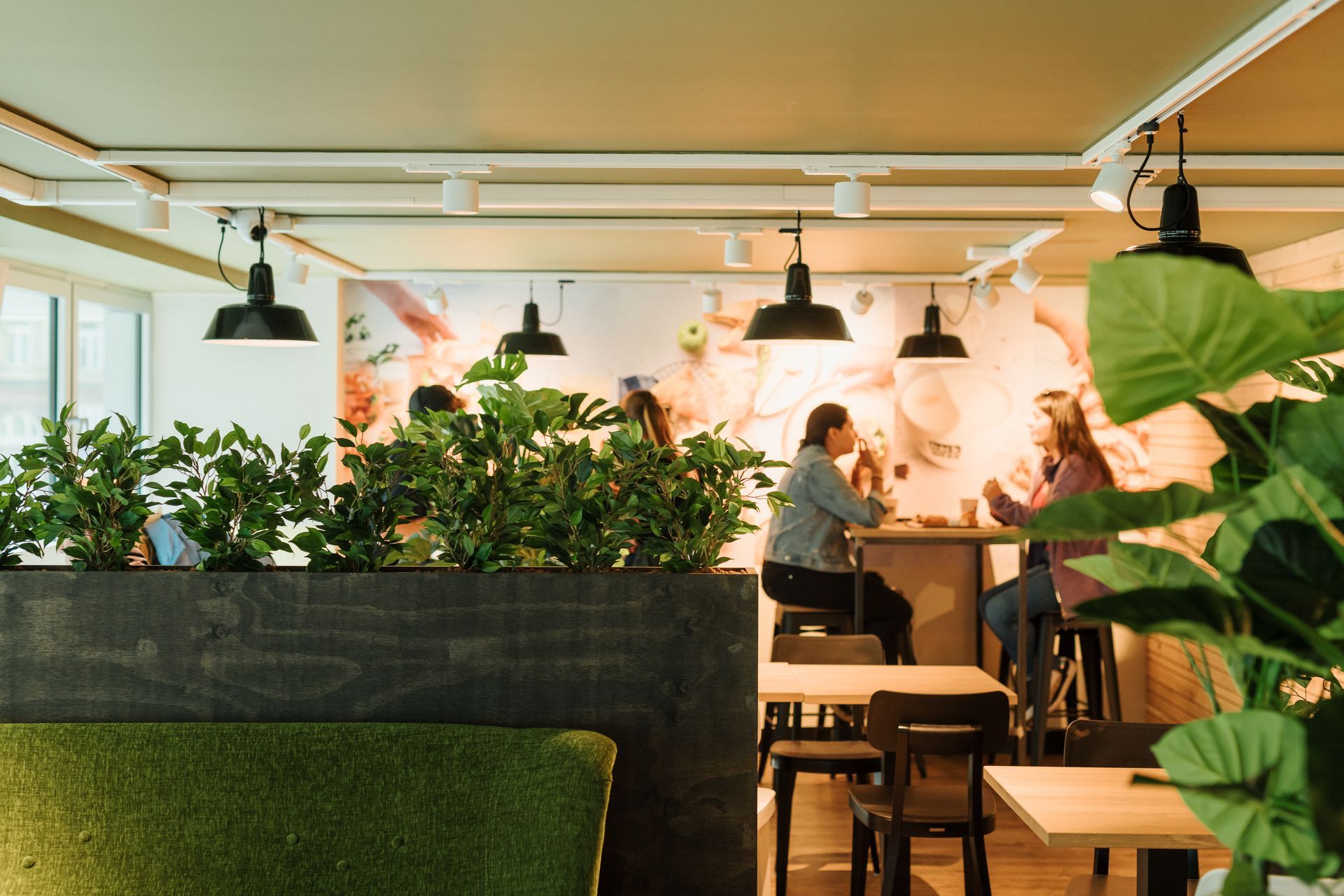 Gezellig café-interieur met planten, hanglampen, tafels en een muur bedekt met een muurschildering; iemand zit aan de bar.