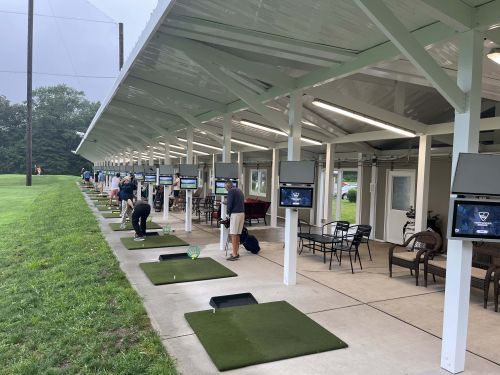 Golf driving range with players hitting balls. Covered stalls with screens and grass areas.