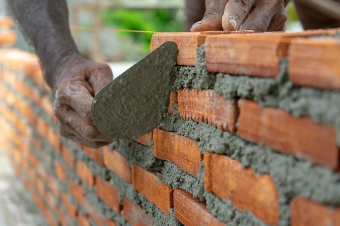 Hands laying gray mortar between orange brick wall courses, close-up construction work