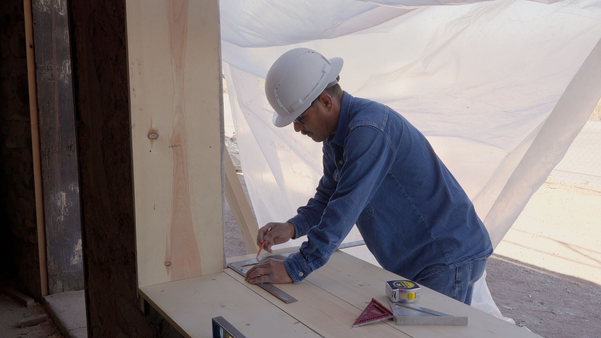 Construction worker in a hard hat measures and marks wood near a window, outdoors under a tarp.