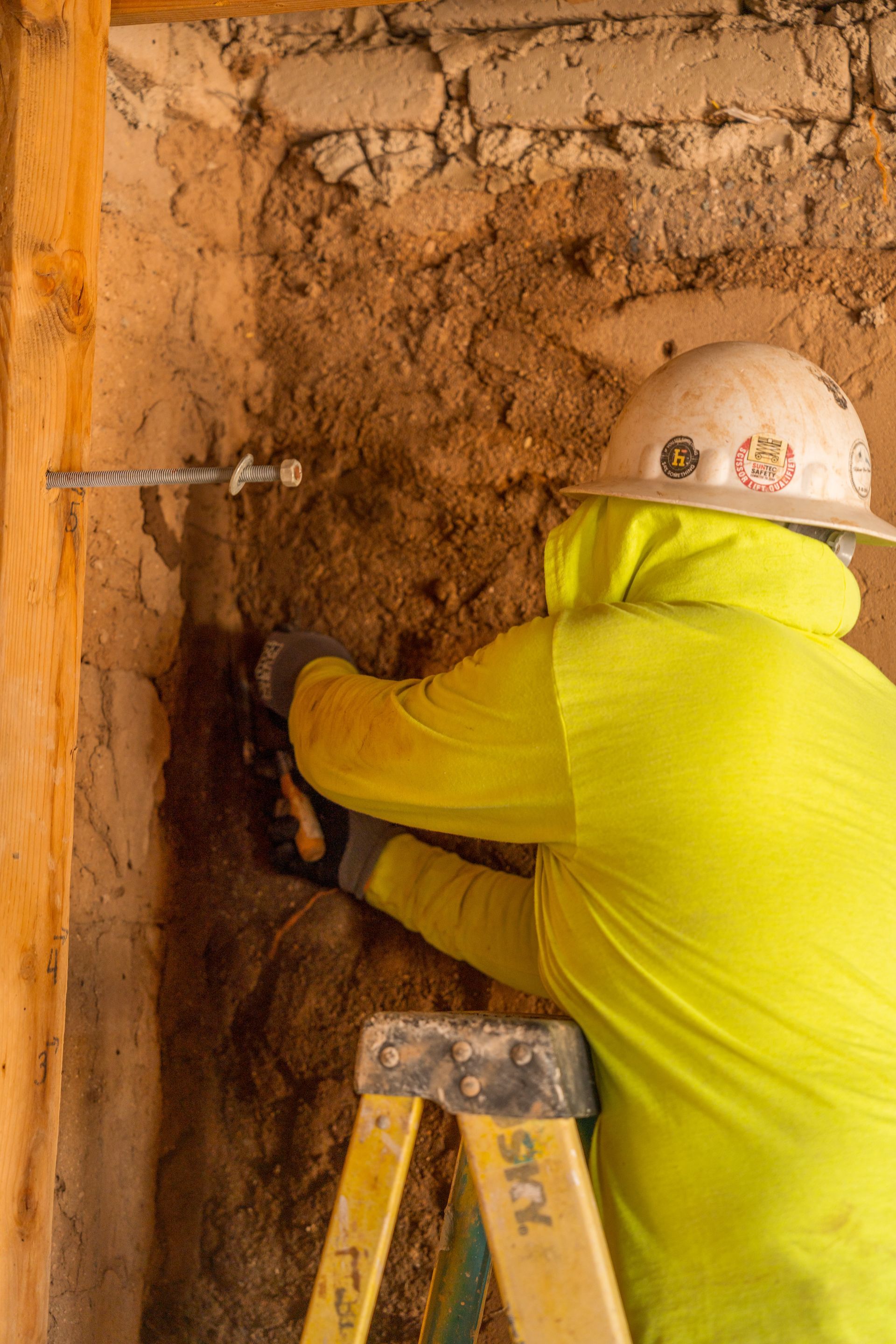 Construction worker in yellow, drilling into a wall; wood framing visible.
