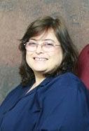 A woman wearing glasses and a blue shirt is smiling for the camera.