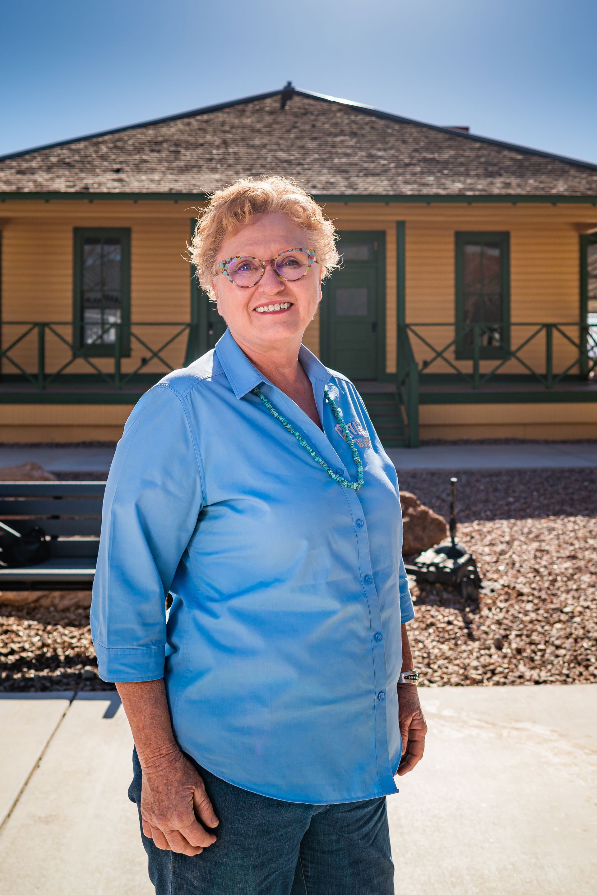 A woman in a blue shirt and glasses is standing in front of a yellow building.