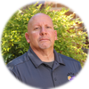 A bald man in a black shirt is standing in a circle in front of a bush.