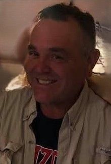 A man in a tan shirt and a black shirt is smiling.