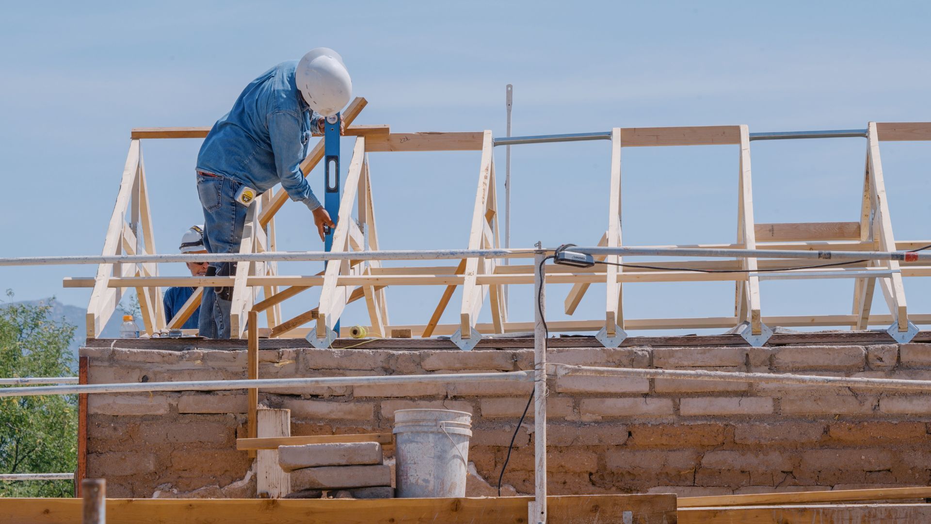 Construction worker using a level on wooden frame of building under construction; bricks, scaffolding, and clear sky in background.