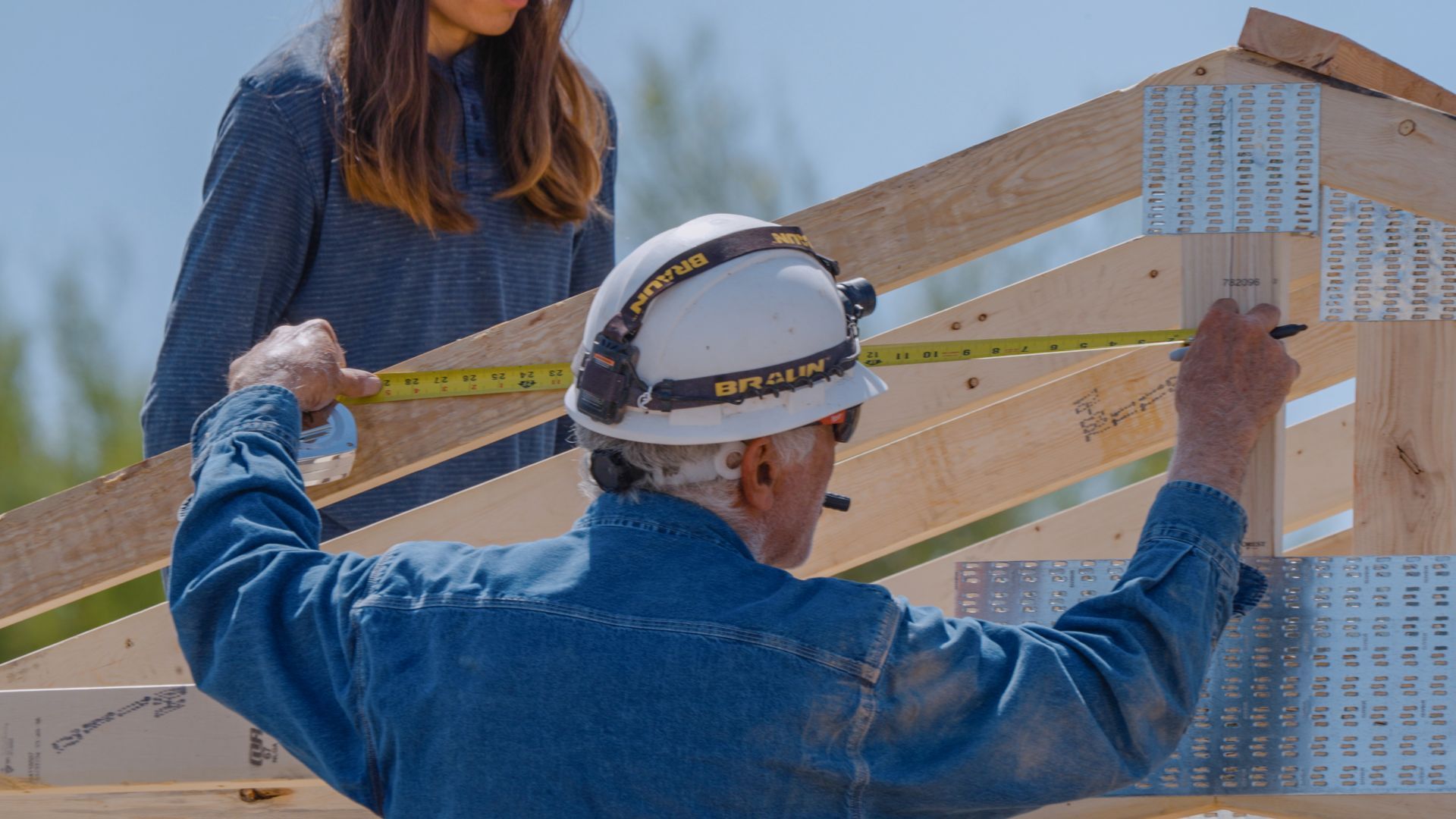 Person in hard hat measures wooden frame with measuring tape; another person watches.