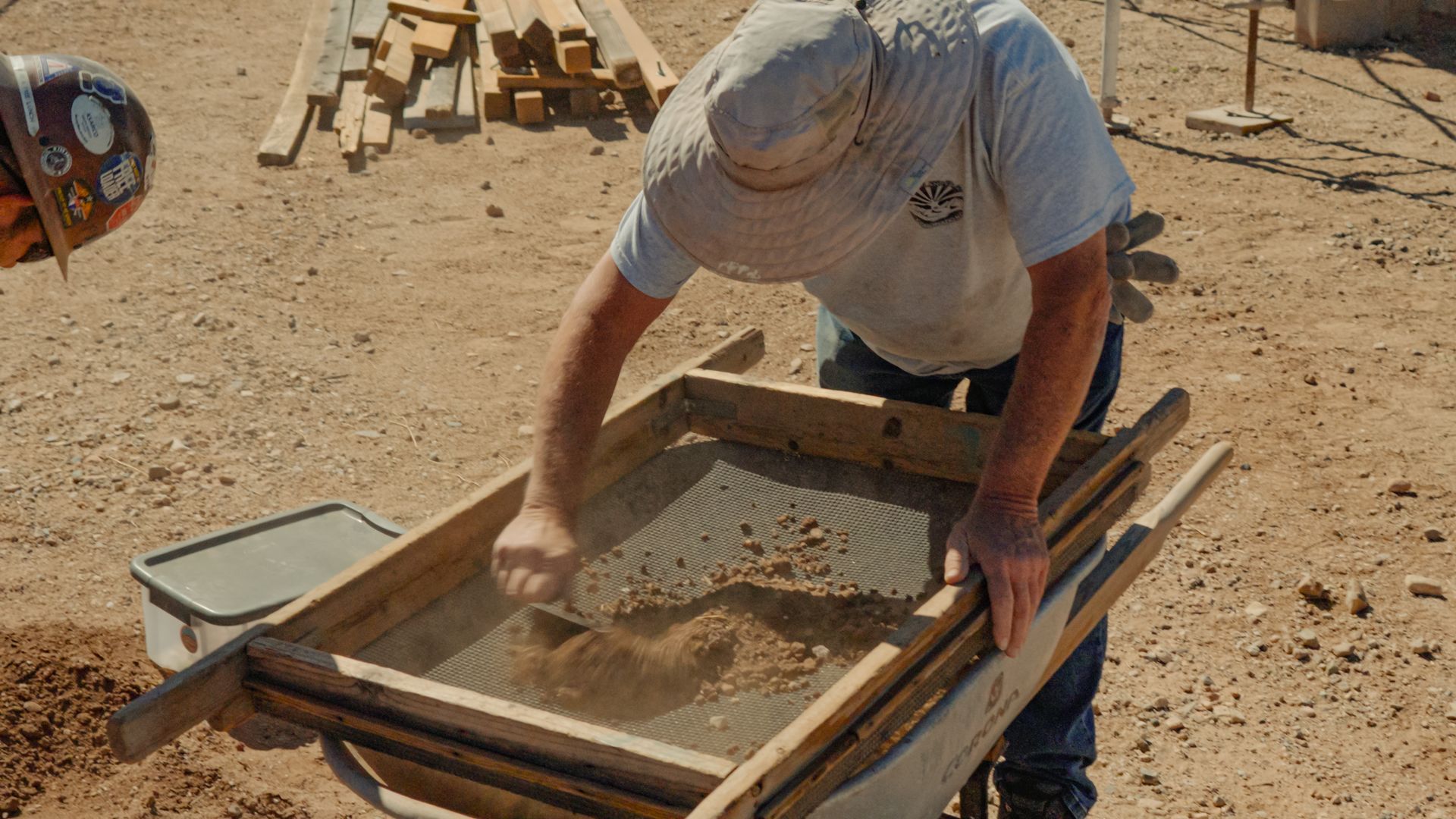 Man sifting dirt with a screen in a dry, outdoor setting, possibly for construction or archaeological work.