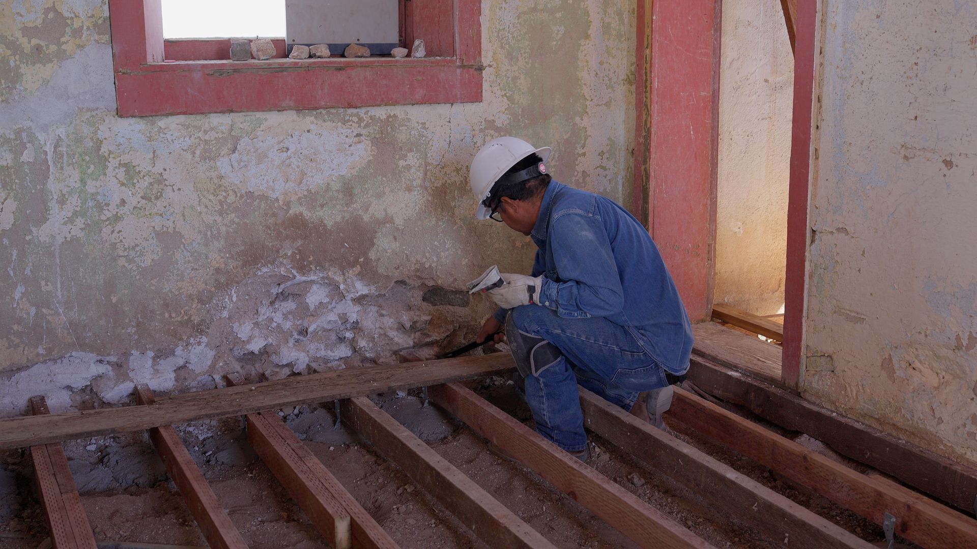 Construction worker inspects exposed wall insulation in a room with a window, wearing a hard hat and blue jumpsuit.