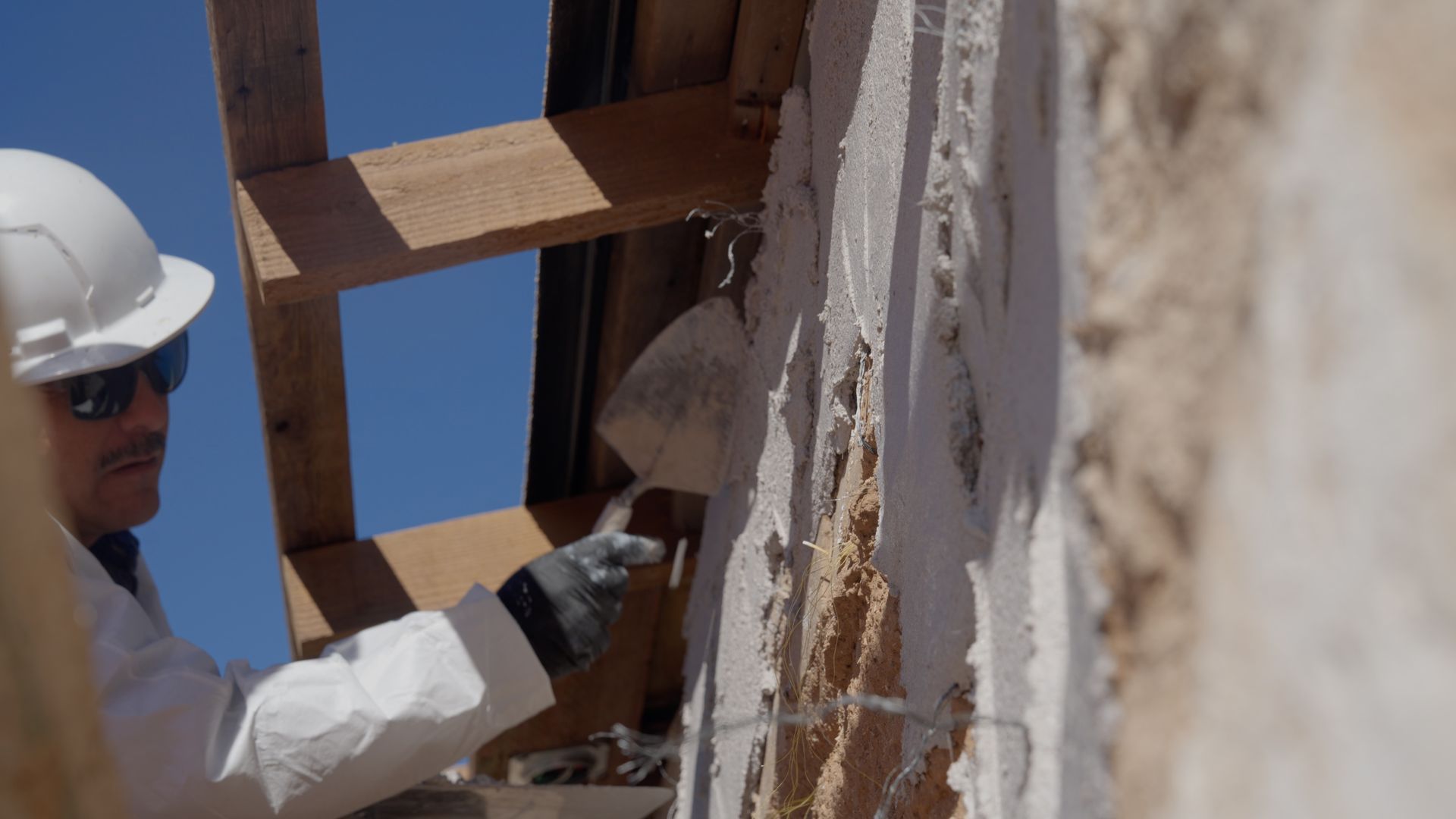Construction worker applying plaster to a wall with a trowel, outdoors in sunlight.