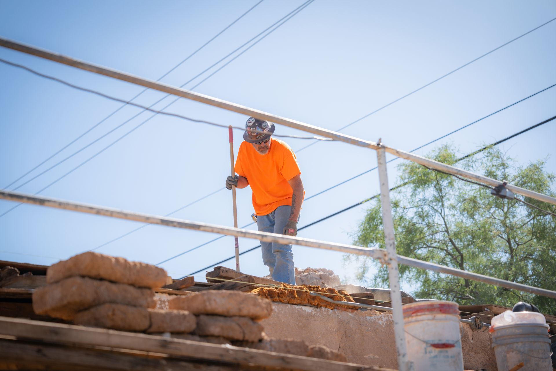 A man in an orange shirt is standing on a roof holding a shovel.