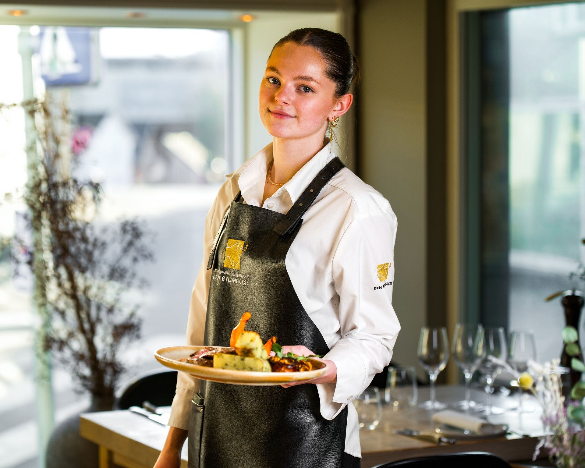 Kvinde i hvid kokkekittel og sort forklæde holder en tallerken mad på en restaurant.