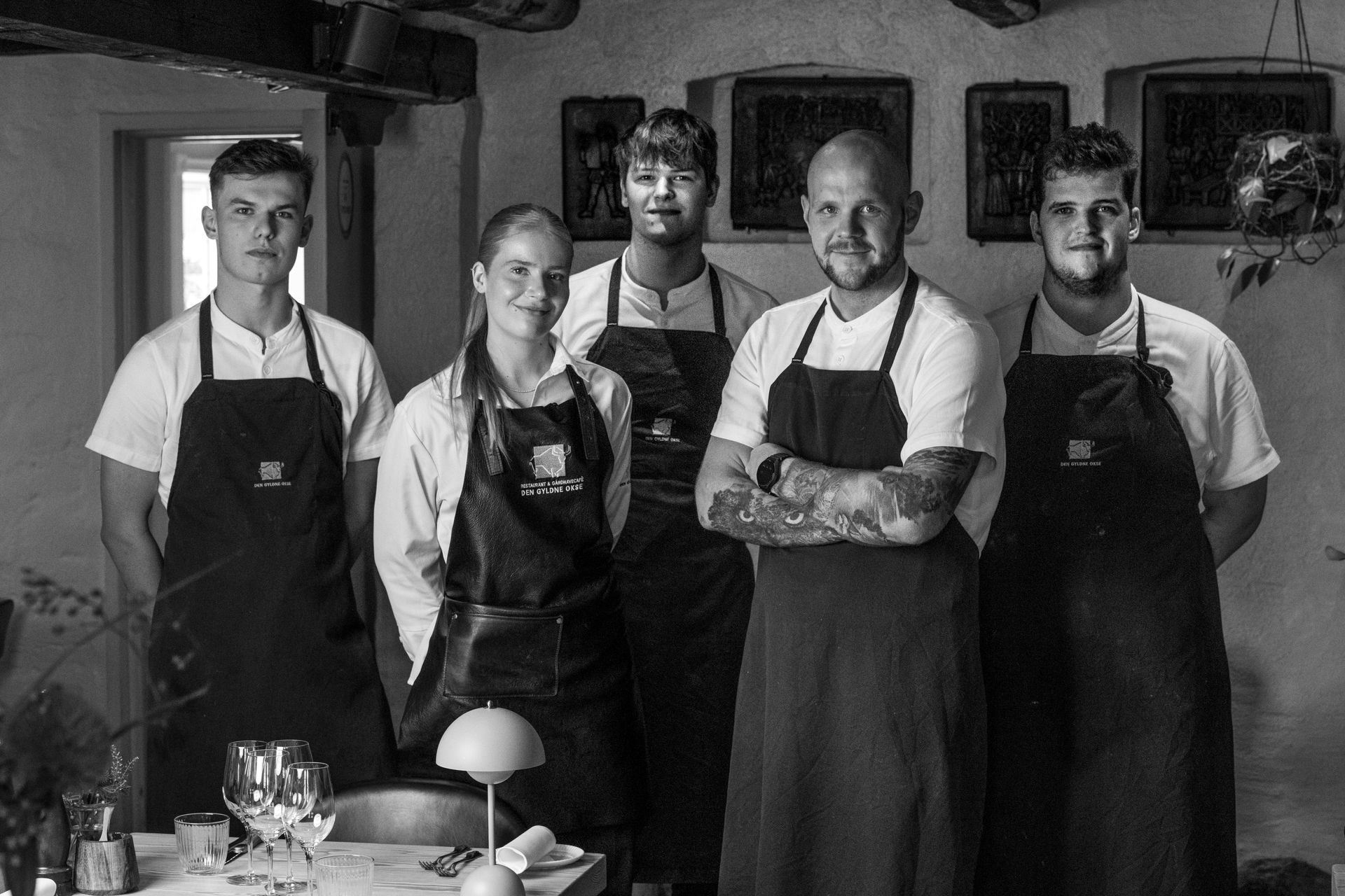 Fem restaurantansatte i sorte forklæder, med armene over kors, i et rustikt køkken.